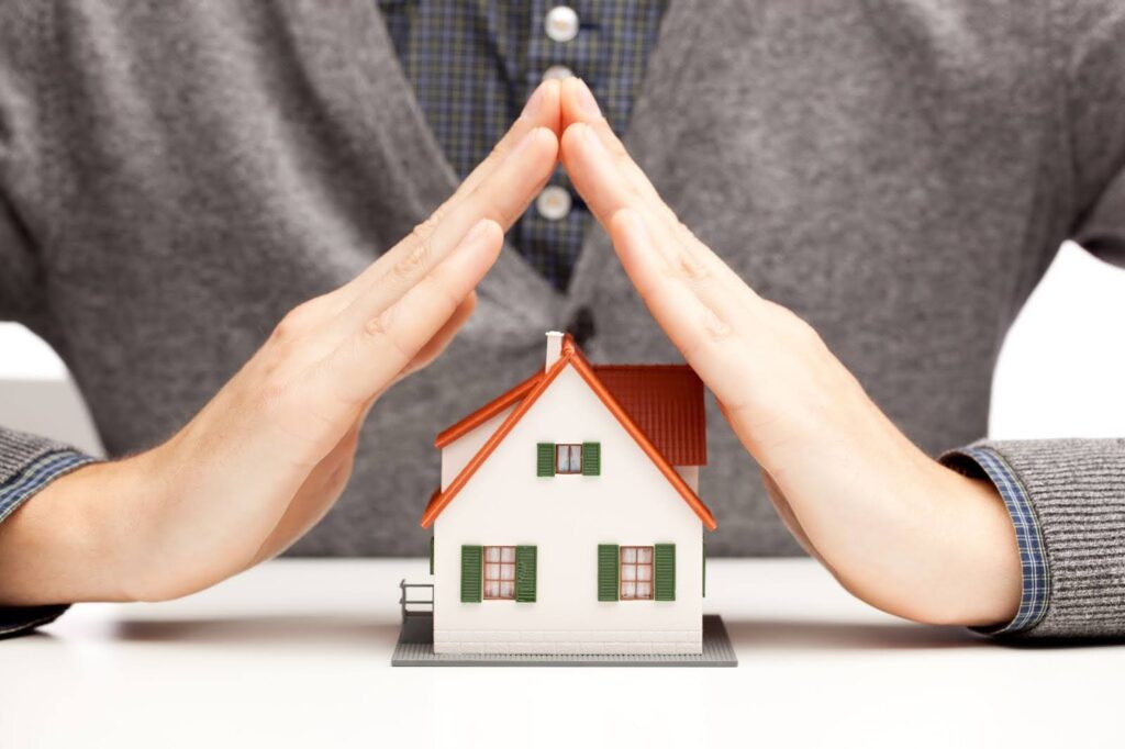 A man sitting behind a toy house with his hands tented over the home to symbolize insurance protection
